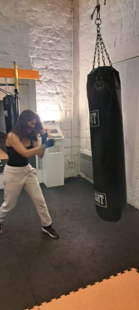 Entraînement de Boxe Femme : Session avec Sac de Frappe Femme s'entraînant avec un sac de frappe noir marqué HT 1963 dans une salle de boxe aux murs de pierre blanche.
