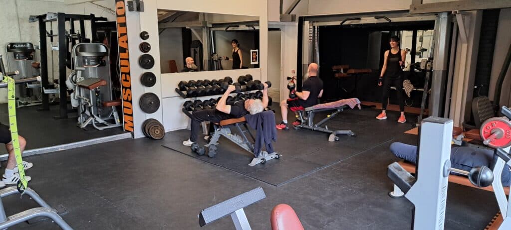 Entraînement en Salle de Musculation avec Poids et Haltères Séance de sport en salle de musculation avec haltères, bancs et machines. Texte MUSCULATION visible sur un pilier.